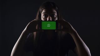 Woman in darkly lit background holds her phone with the WhatsApp logo on the screen in front of her mouth