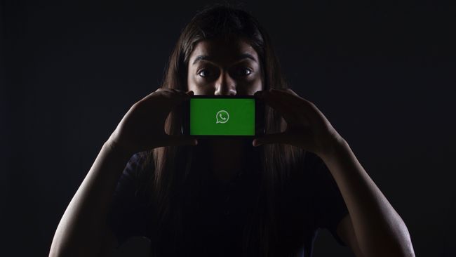 Woman in darkly lit background holds her phone with the WhatsApp logo on the screen in front of her mouth
