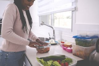 A woman is in the kitchen, she is filling containers with left over food including vegetables and fruit.