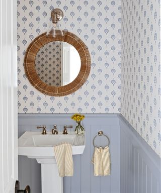 Farmshouse style with pastel blue wainscotting, ditsy patterned wallpaper, yellow and white stripe towels, rattan mirror