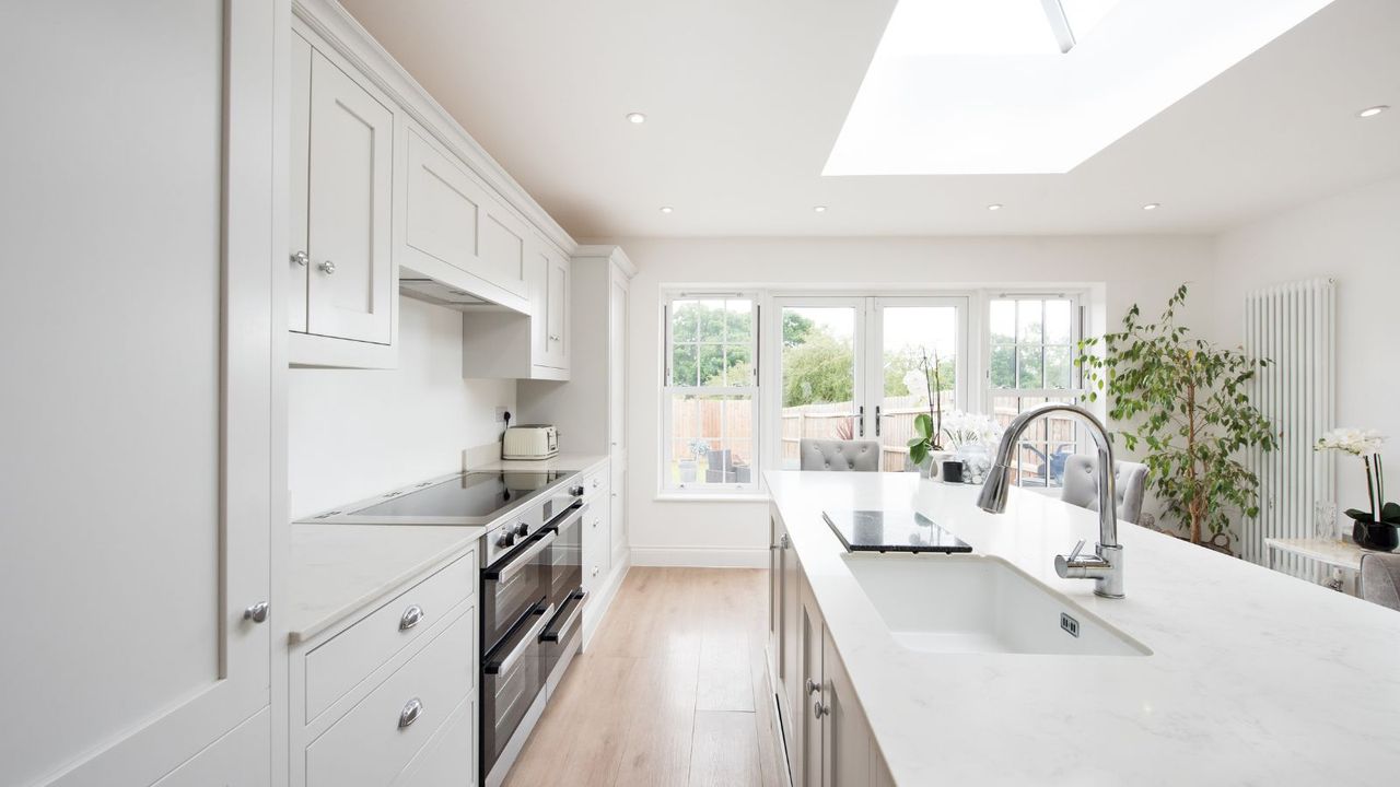 A general interior view of a domestic dove grey kitchen, with painted white walls, island with quartz worktop, chrome tap, grey crushed velvet bar stools, stainless steel range cooker oven, chrome tap, skylight, vertical radiator, potted plant and patio doors into the back garden within a home