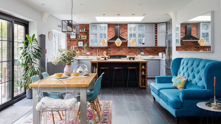 Open-plan kitchen diner with grey Shaker-style kitchen and wooden island, brick slip wall, wooden dining table and chairs on a pink patterned rug, and a large blue velvet sofa