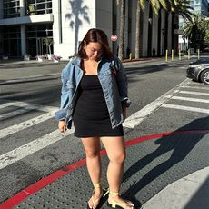 marina wears black mini dress, denim jacket and butter yellow sandals while standing on a street corner