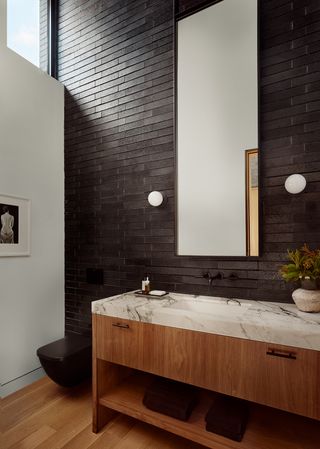 A bathroom vanity with a dark tiled wall and a large mirror. a large clerestory window is overhe