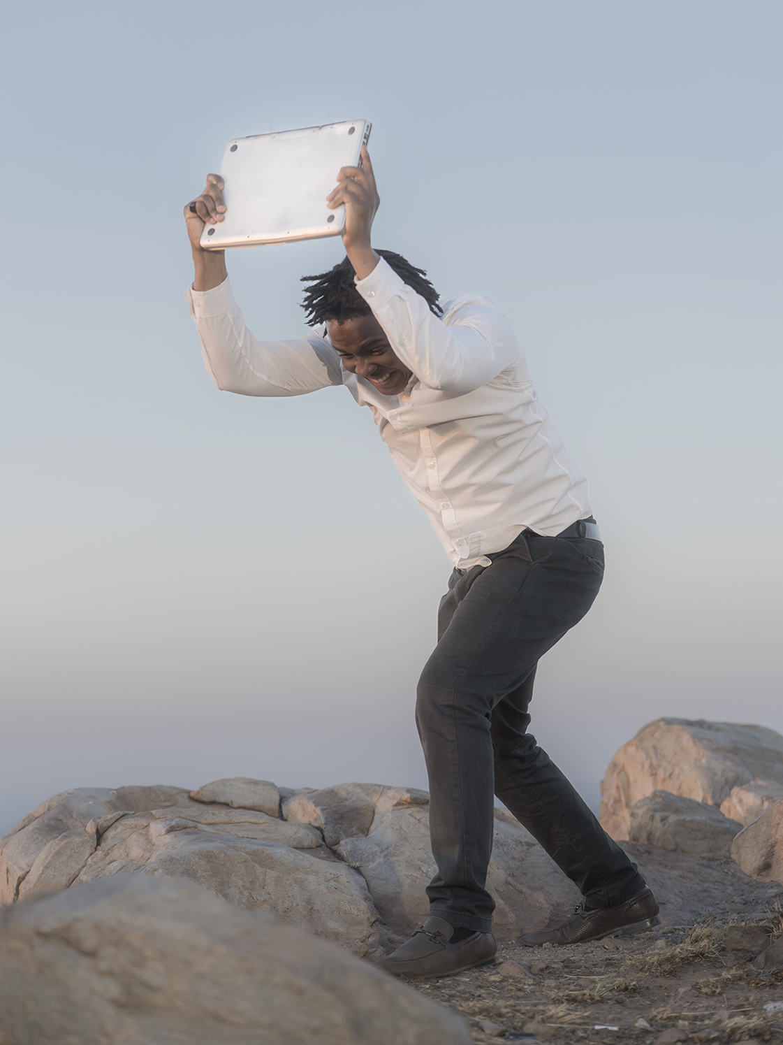 Colour film still of a man holding a laptopraised above his head with the aim of hitting it against therocky ground that he is standing on.