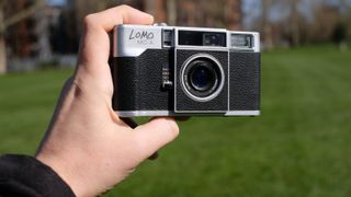 Photograph of a man's hand holding the Lomography Lomo MC-A analog compact camera, front facing