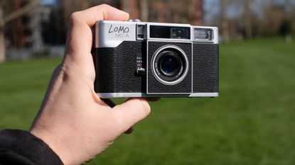 Photograph of a man's hand holding the Lomography Lomo MC-A analog compact camera, front facing