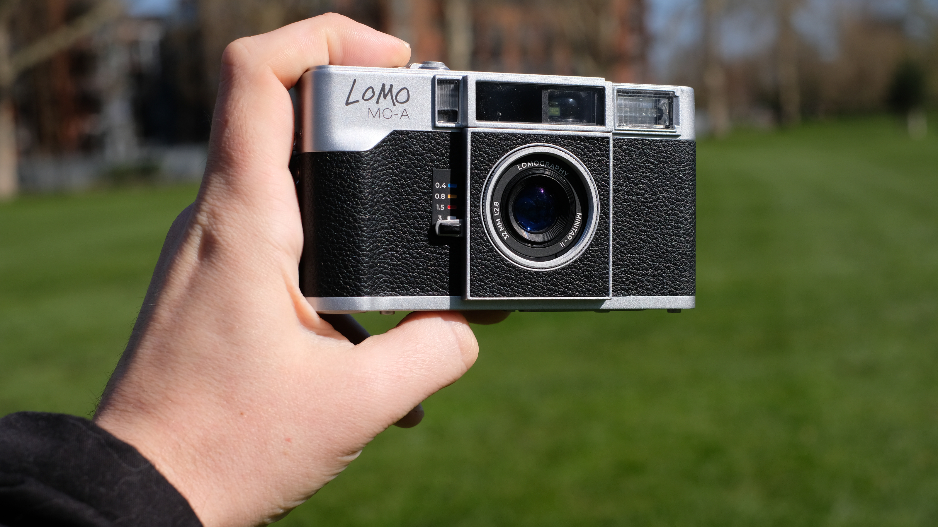 Photograph of a man's hand holding the Lomography Lomo MC-A analog compact camera, front facing