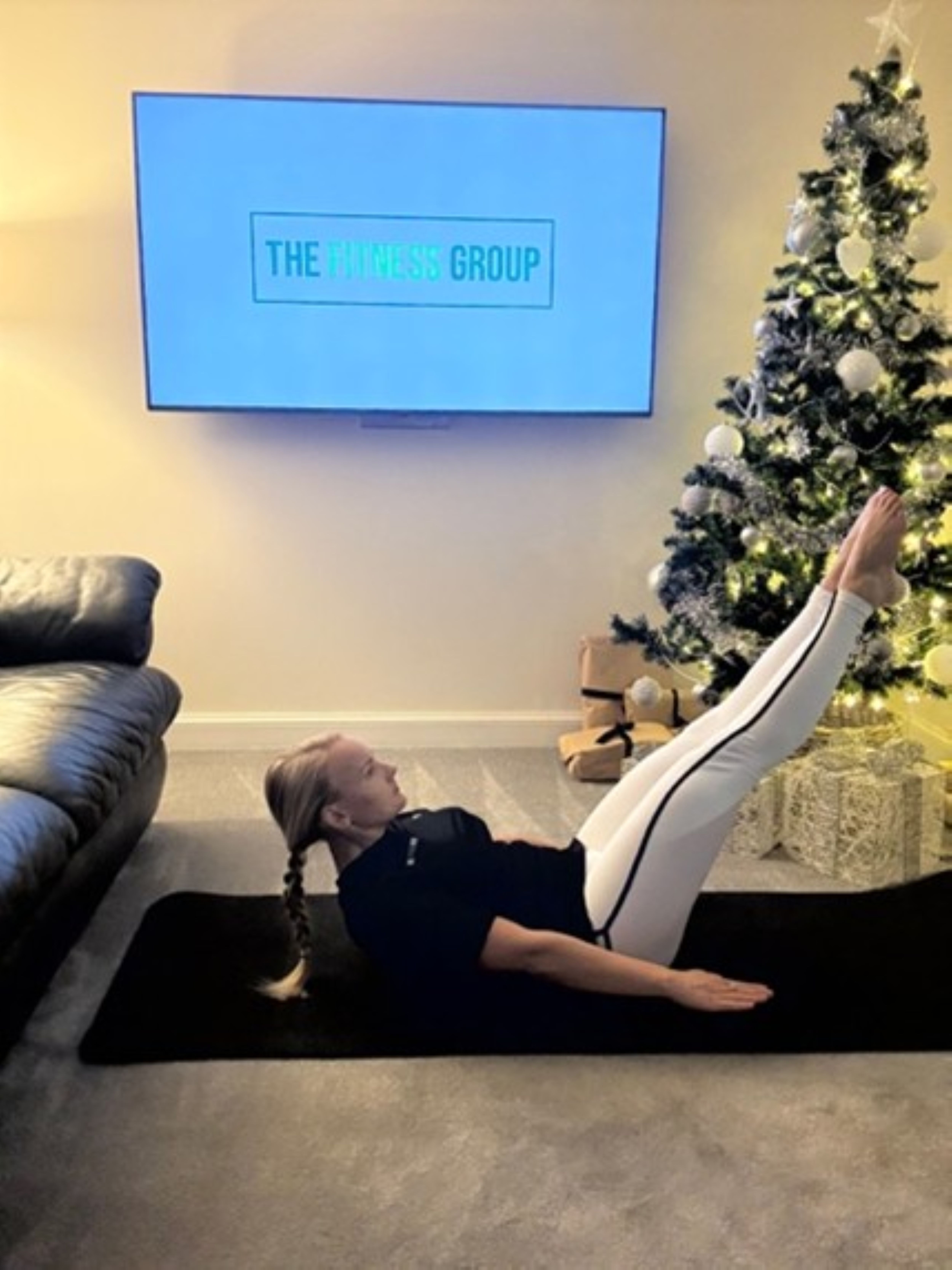 Woman demonstrates a Pilates exercise between a sofa and a Christmas tree. A mounted TV in the background reads "The Fitness Group"