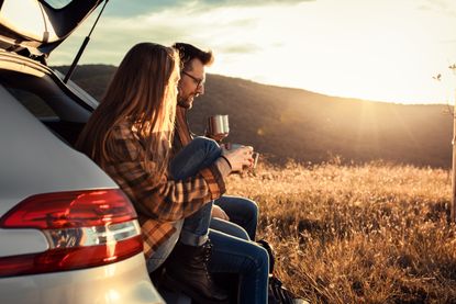 Couple on road trip sitting in boot of a car drinking coffee