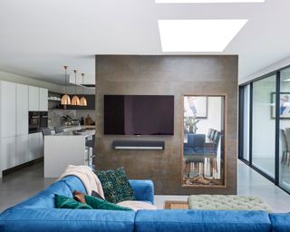 An example of broken plan living room ideas showing a blue corner sofa in front of a partition featuring a glass-walled fireplace and a TV