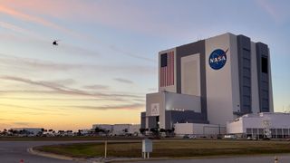 A helicopter flies by NASA's tall, rectangular Vehicle Assembly Building as a sunset-kissed sky fades from blue to hues of orange and yellow behind wisps of pink-tinted clouds.