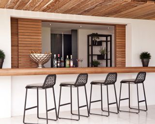 Garden bar ideas featuring a long wooden bar with black bar stools in a white painted outdoor structure.