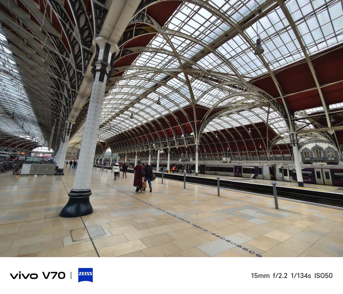 Wide-angle view along railway platform with arched glass roof, passengers walking and train at platform.