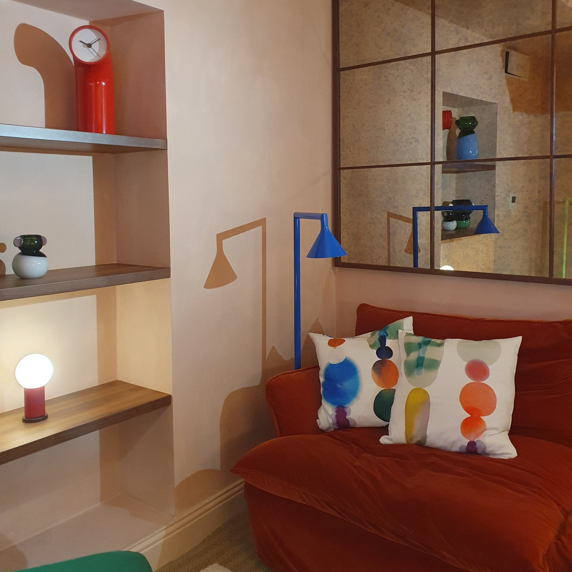 A living room set up with pieces from the IKEA PS 2026 collection including the floor lamp in blue positioned as a reading light and the red table clock displayed on a shelf