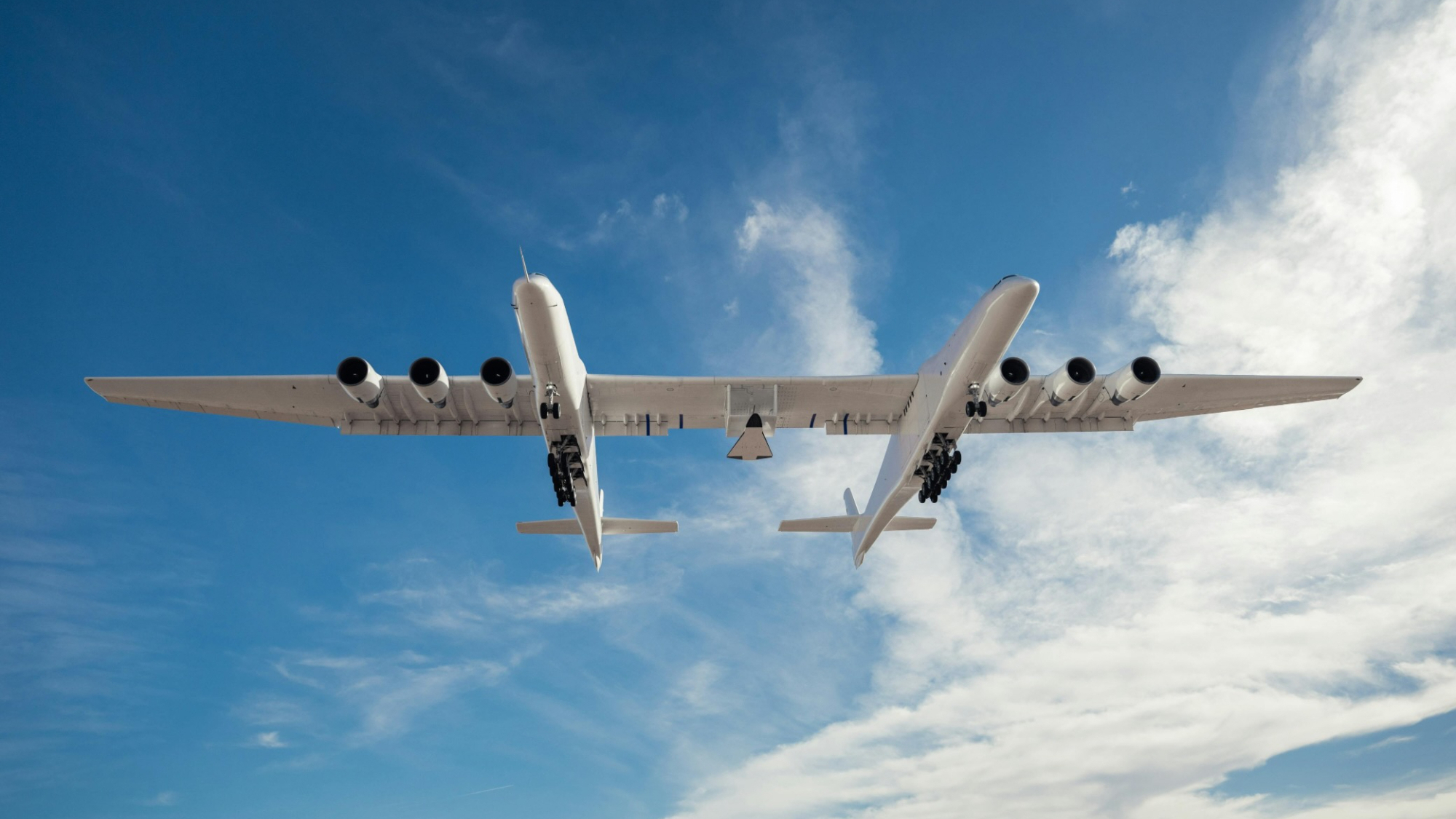 Vista de um avião branco de transportadora de dupla fuselagem em voo de baixo, com um céu azul nublado ao seu redor
