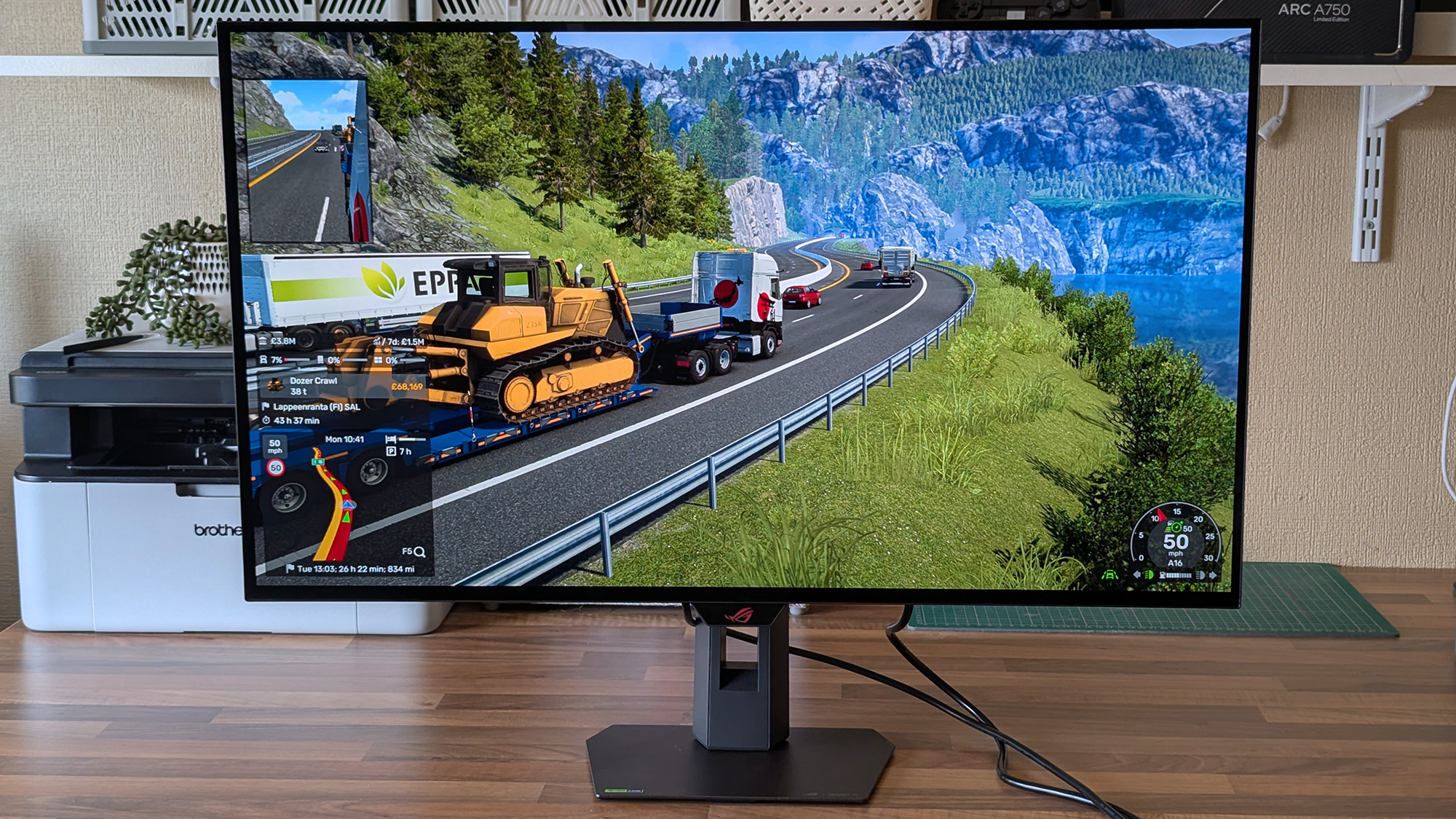 ASUS ROG Strix OLED XG32UQWMS monitor shows Euro Truck Simulator 2 hauling a yellow bulldozer along a mountain road, with delivery details and route information visible on&amp;#8209;screen.