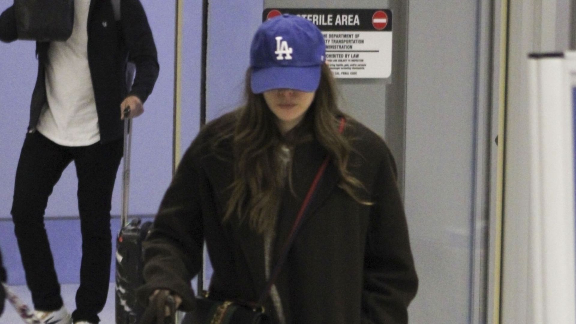 Elizabeth Olsen walks through the airport wearing black leather ankle boots, a black wool coat and a blue cap.