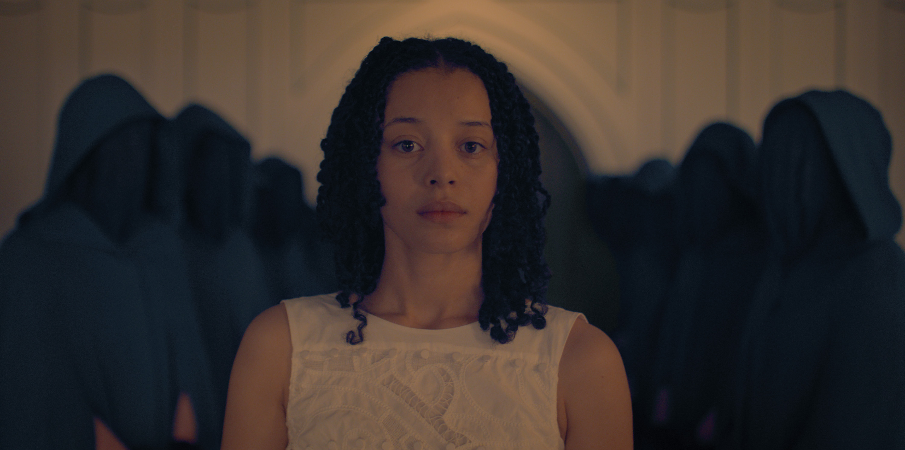 A young woman (Chase Infiniti) wears a white top while standing in a room filled with people, their faces obstructed by black robes. A first-look image from Hulu's 'The Testaments.'