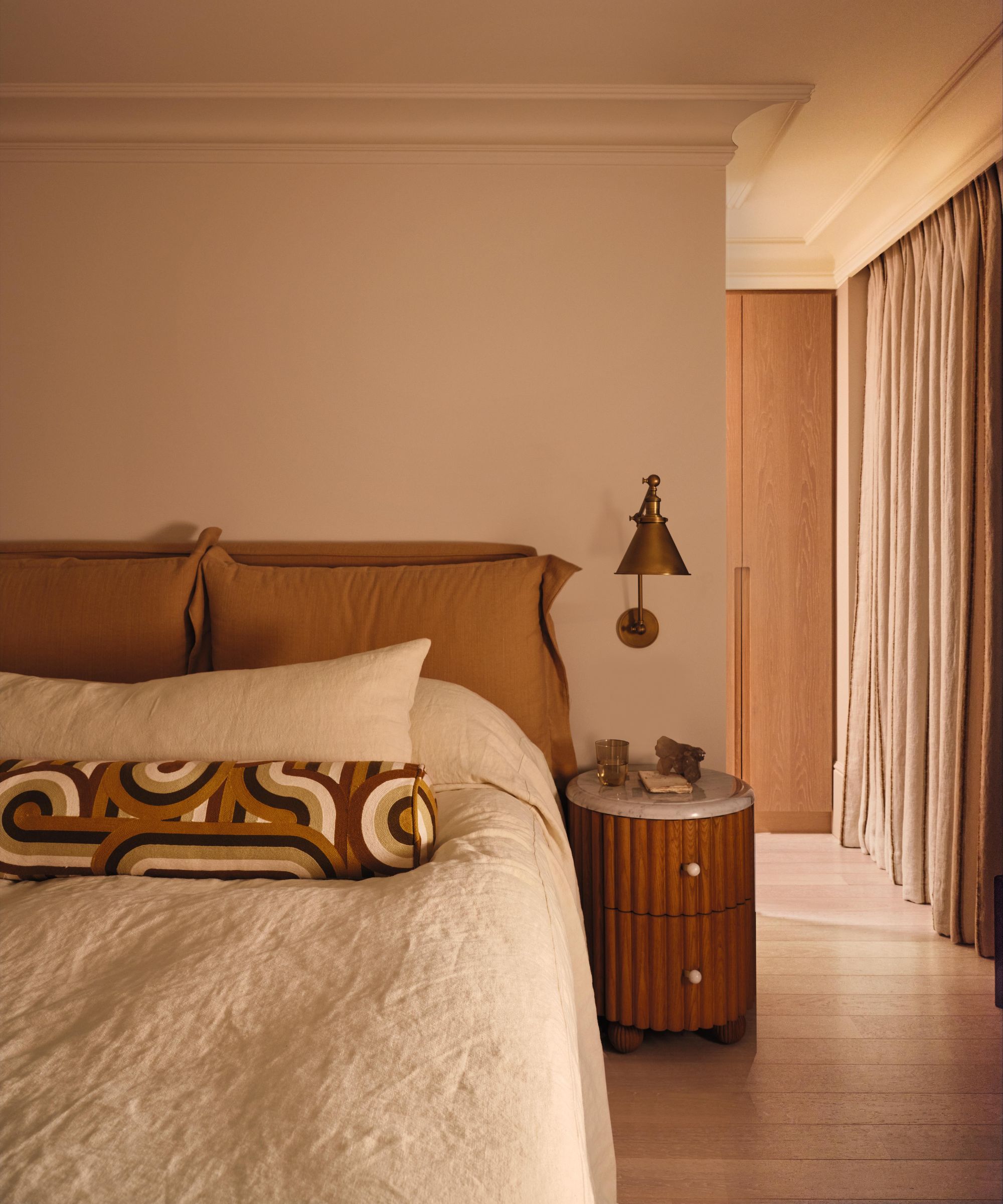a neutral wabi-sabi bedroom with a mustard pillow back headboard, soho home wooden nightstand and brass sconce