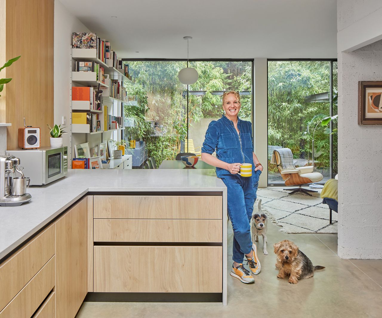 Woman in blue jumpsuit casually leaning against a kitchen peninsula in an open plan kitchen and dining space in a contemporary home renovation. Two small dogs are at her feet