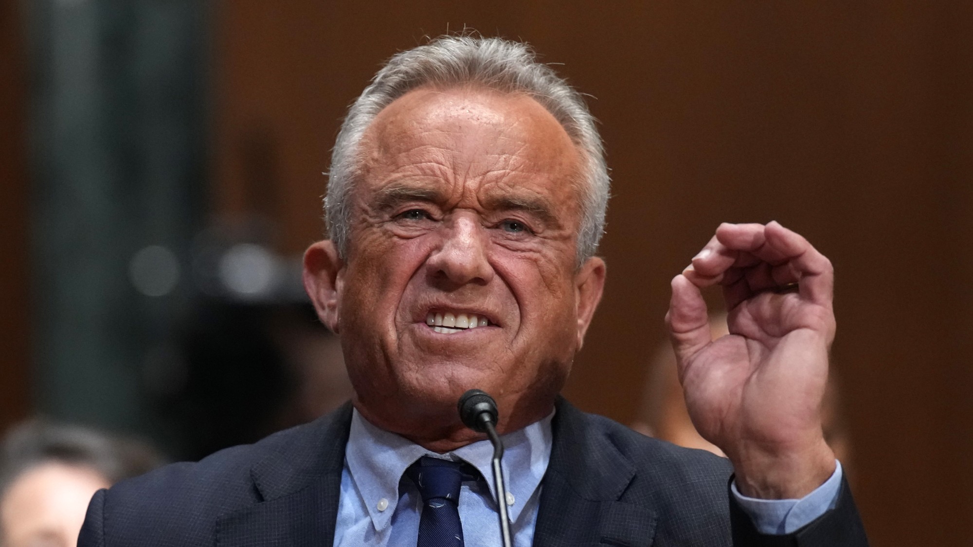 Robert F. Kennedy JR, grimacing and holding hand up in front of a mic at a hearing