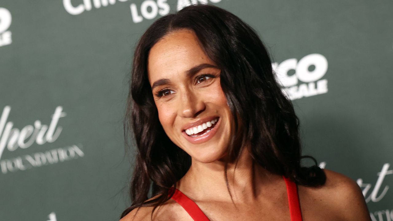 Meghan Markle wearing a red sleeveless dress and smiling in front of a step and repeat