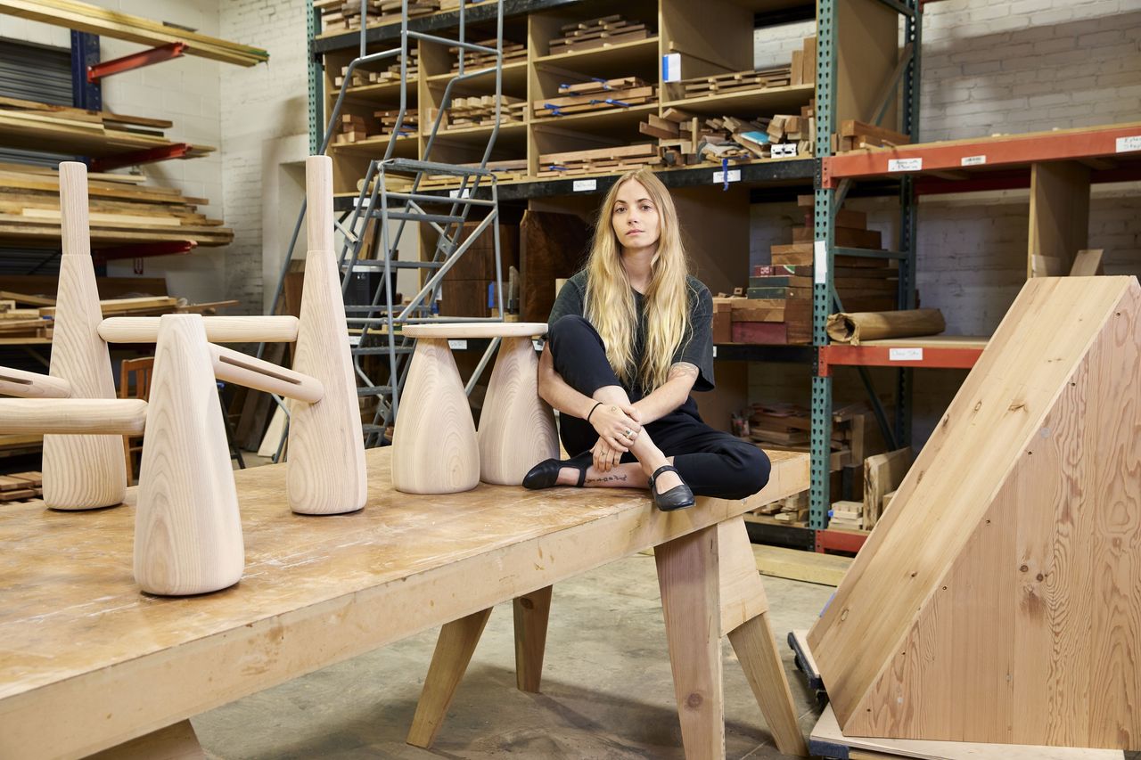 A young girl dressed in a grey T-shirt and black trousers sits on a wooden table next to her furniture creations in a workshop.