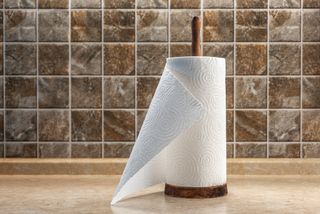 A roll of kitchen roll on a wooden holder on top of a kitchen countertop, and in front of a tiled background.