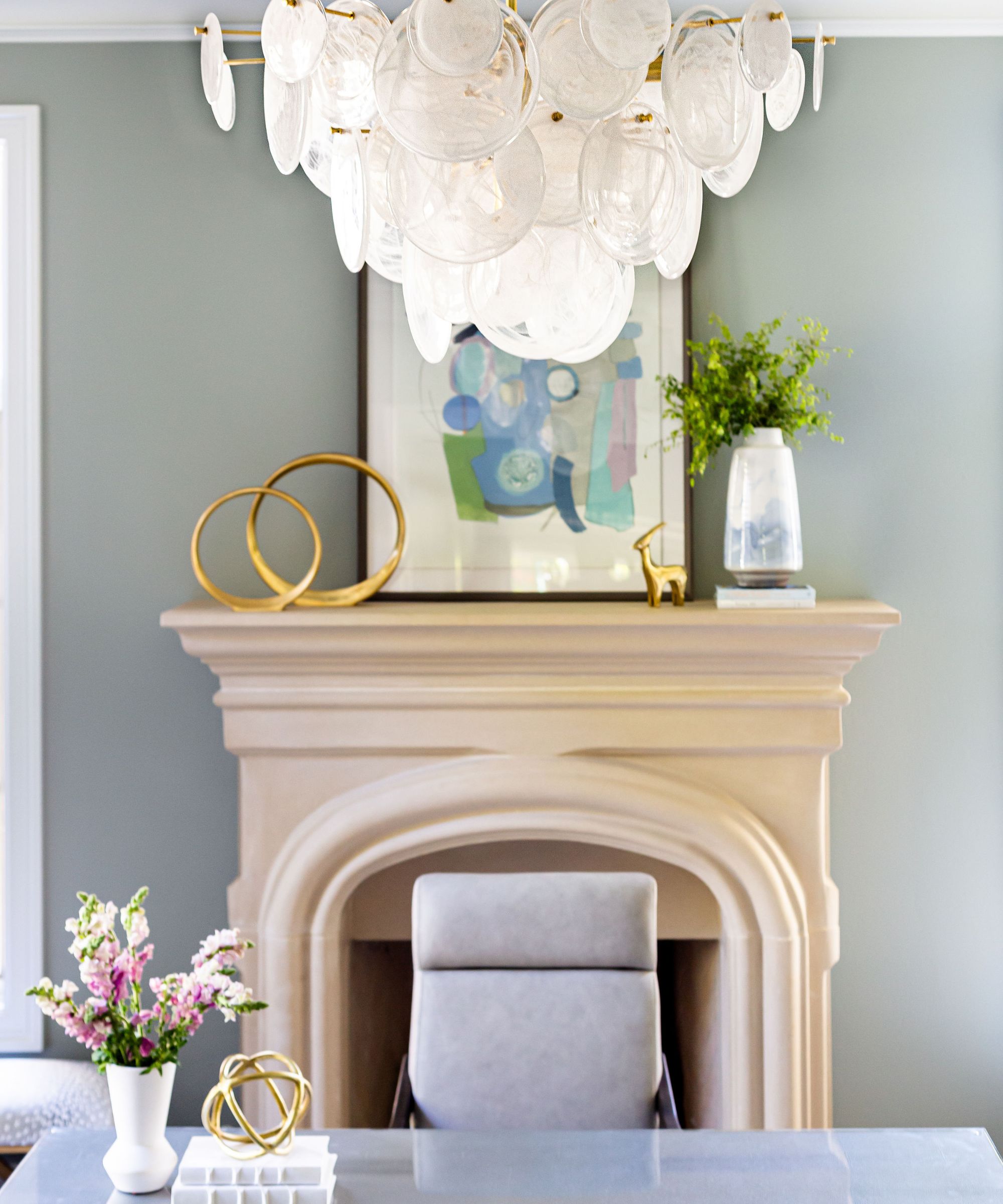 Home office with Oyster Bay by Sherwin-Williams painted walls, a stone fireplace, a glass desk, a grey office chair, and an abstract print on the mantel with a vase of foliage and gold statues.
