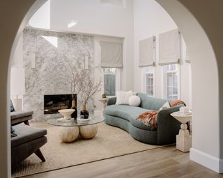 Living room through large archway with white walls, light wood floors, beige rug, sculptural coffee table, light teal sofa and large white marble fireplace