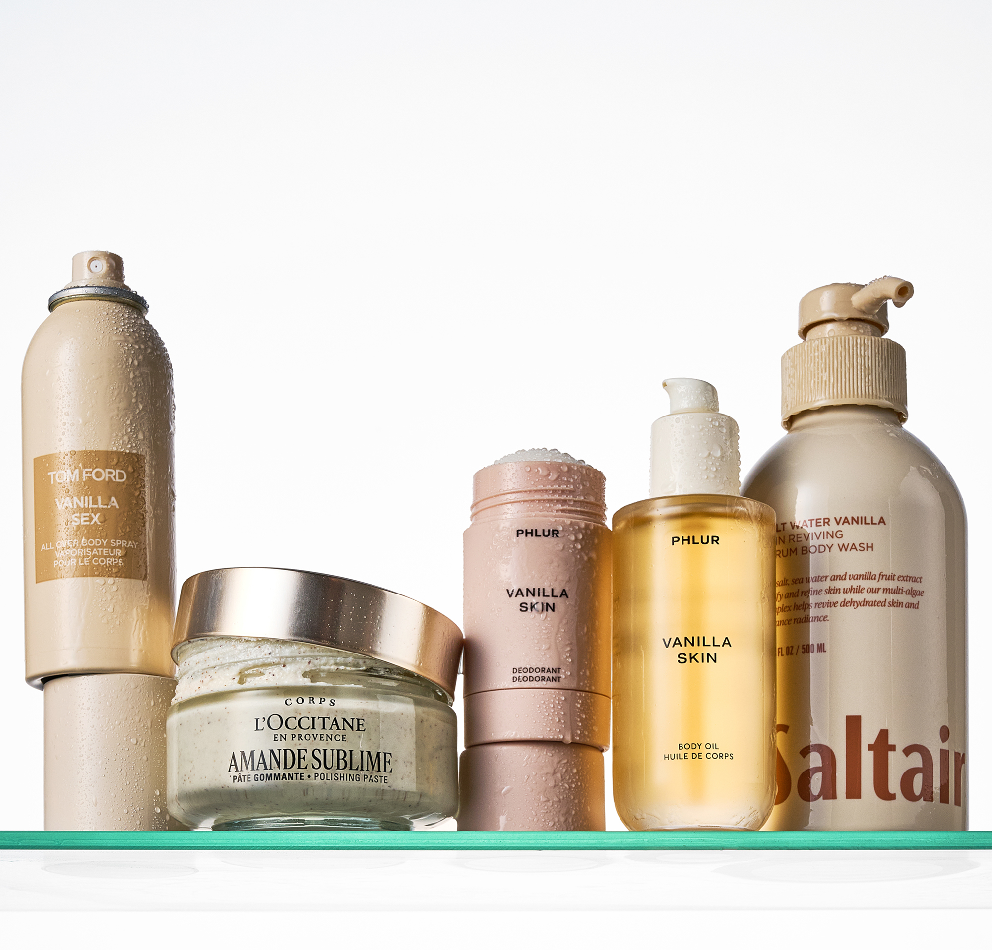A picture of a Tom Ford body spray, a L'Occitane body scrub, a Phlur deodorant and body oil and a Saltair body wash on a shower shelf