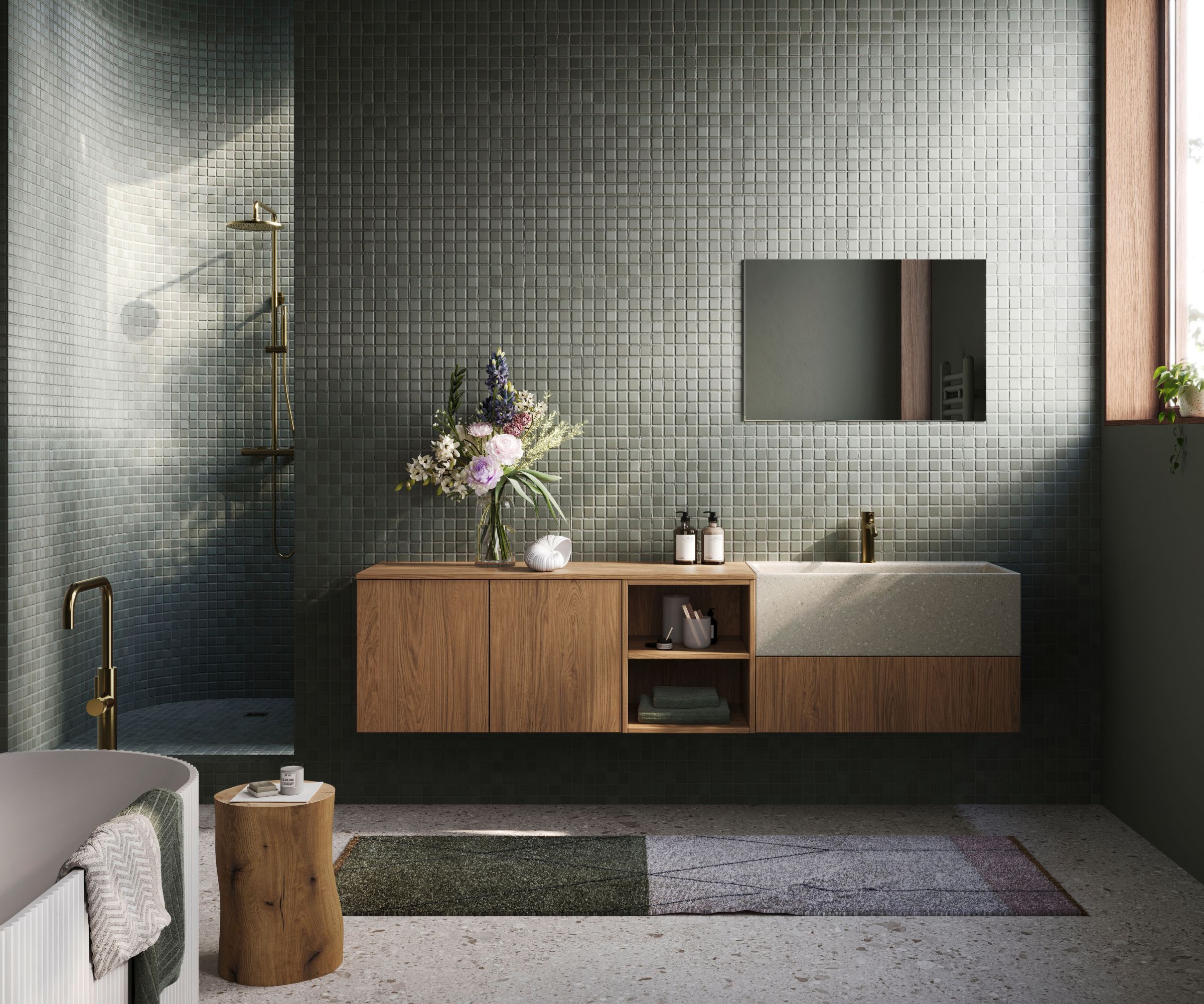 A dramatic bathroom with floor-to-ceiling dark green mosaic tiles, a floating oak vanity unit with open shelving and a stone basin, and brass fittings throughout. A freestanding white bath, rustic wood stump side table, and fresh floral arrangement complete the earthy, luxurious scheme.