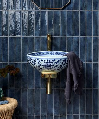 A bathroom featuring deep blue gloss tiles with a reflective finish. A decorative blue and white patterned ceramic sink is mounted on a gold fixture. A folded dark grey towel hangs elegantly from the sink.