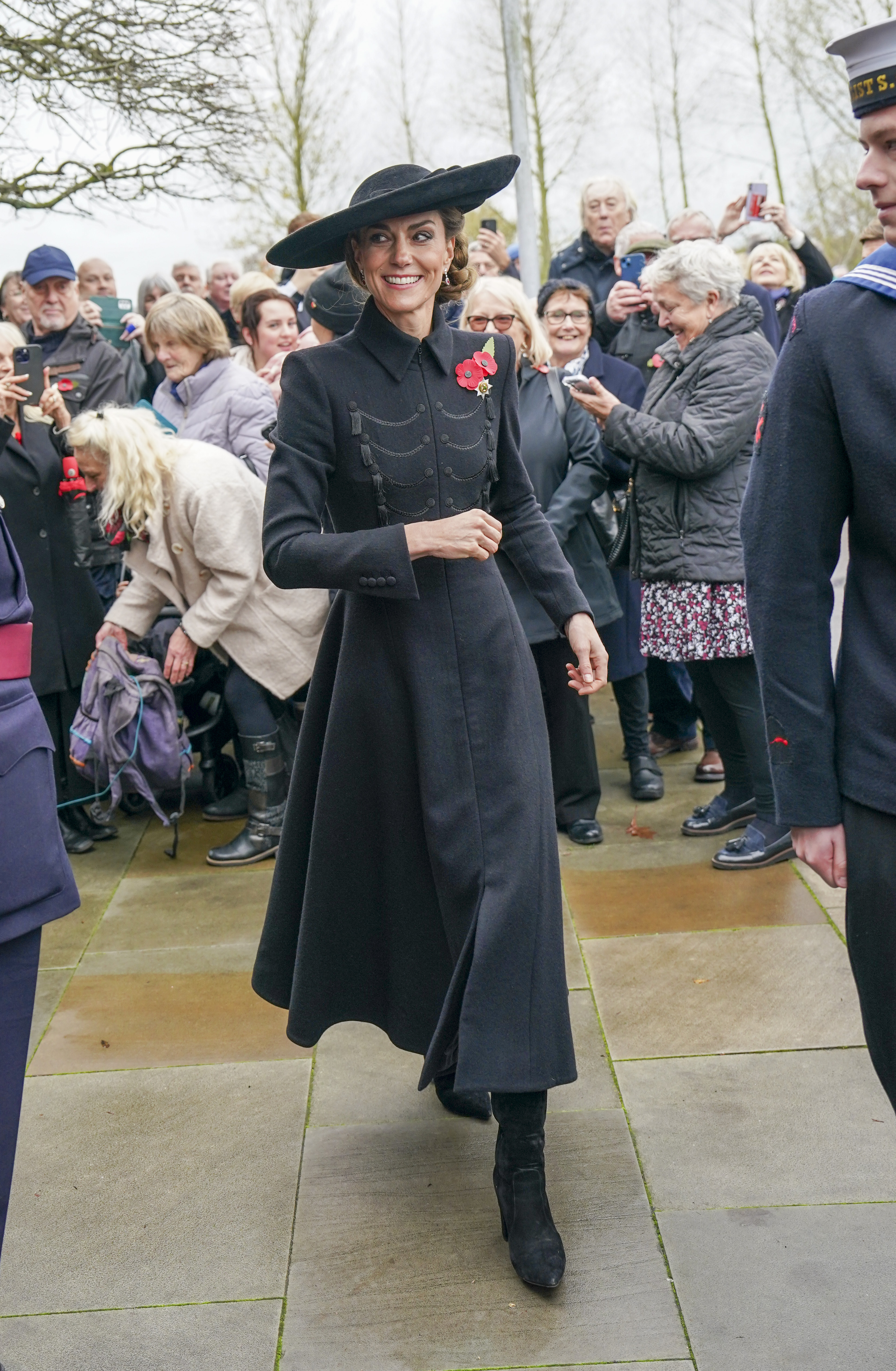 Kate Middleton walking wearing a long black coat and smiling