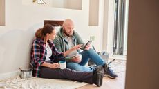 Couple sitting on dust sheets on the floor in newly decorated living room. Both are looking at a mobile phone.