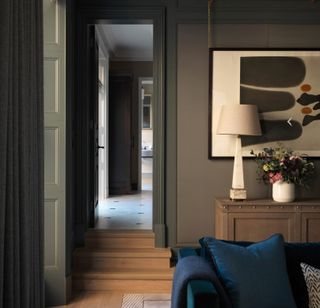View from living room into hallway with dark grey living room walls, wooden flooring and a large piece of abstract art.