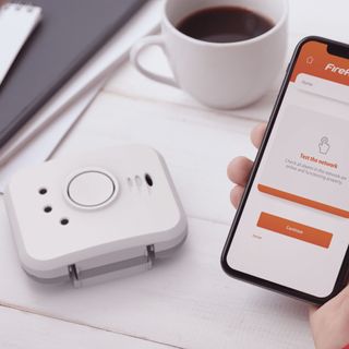 a smart smoke alarm beside a hand holding a smartphone with the companion app on screen