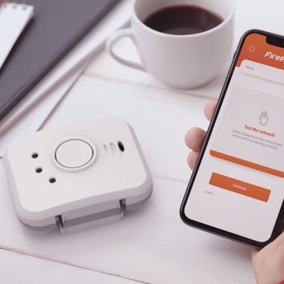 a smart smoke alarms beside a hand holding a smartphone with the companion app on screen