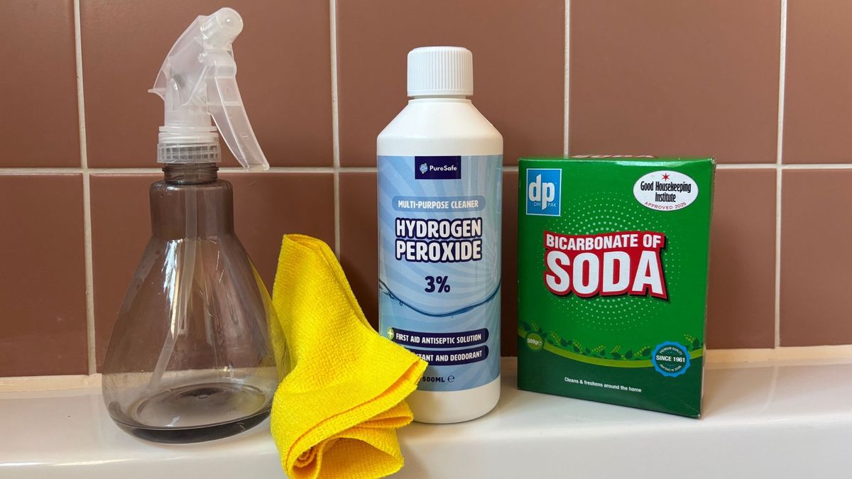 A blue bottle of 3% hydrogen peroxide, green box of baking soda, a yellow microfibre clothe and translucent spray bottle placed on the side of a bath tub, set against orange tiles