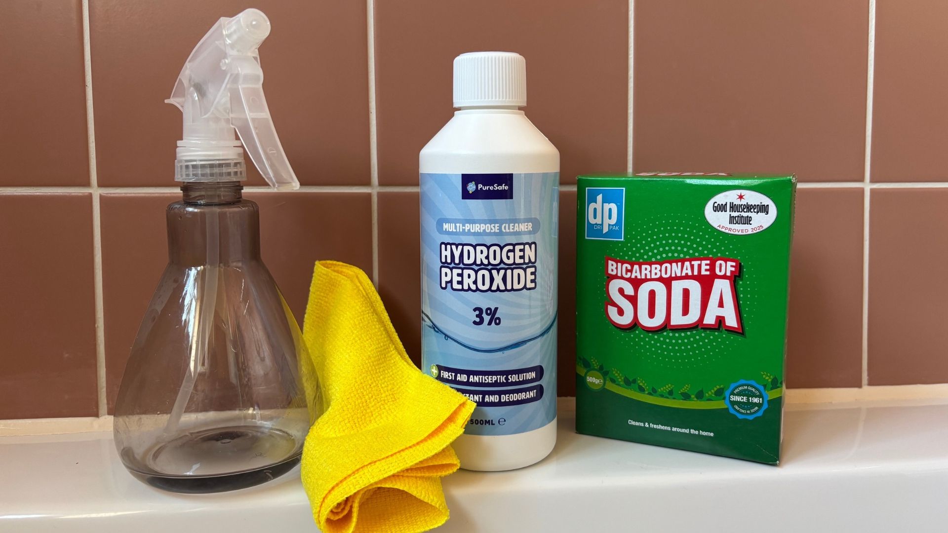 A blue bottle of 3% hydrogen peroxide, green box of baking soda, a yellow microfibre clothe and translucent spray bottle placed on the side of a bath tub, set against orange tiles
