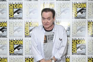 a man in a button-down shirt over a t-shirt smiles for portraits in from of a banner that reads "comic con international"