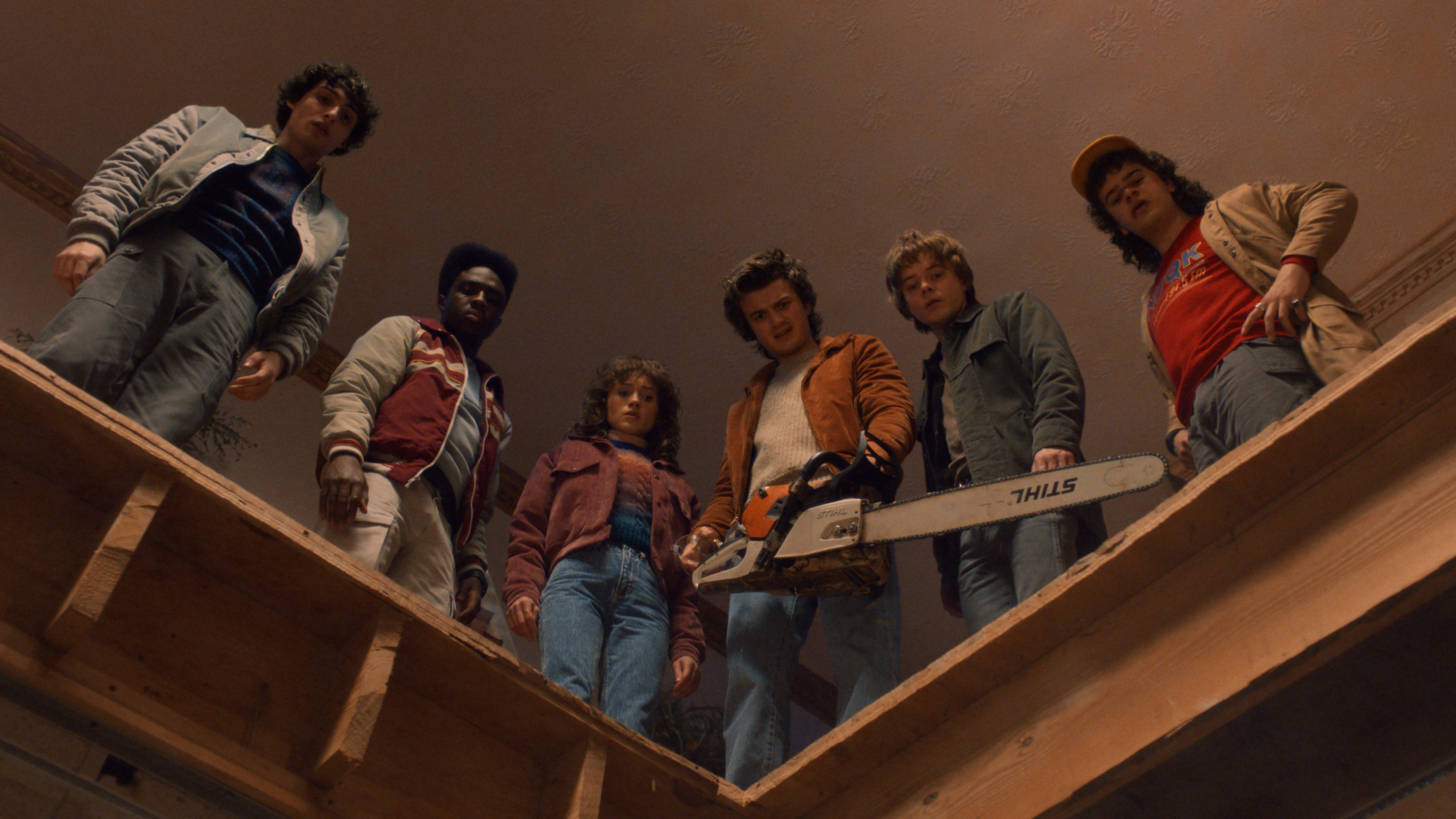 (L to R) Finn Wolfhard as Mike Wheeler, Caleb McLaughlin as Lucas Sinclair, Natalia Dyer as Nancy Wheeler, Joe Keery as Steve Harrington, Charlie Heaton as Jonathan Byers, and Gaten Matarazzo as Dustin Henderson standing on a ledge and looking down.