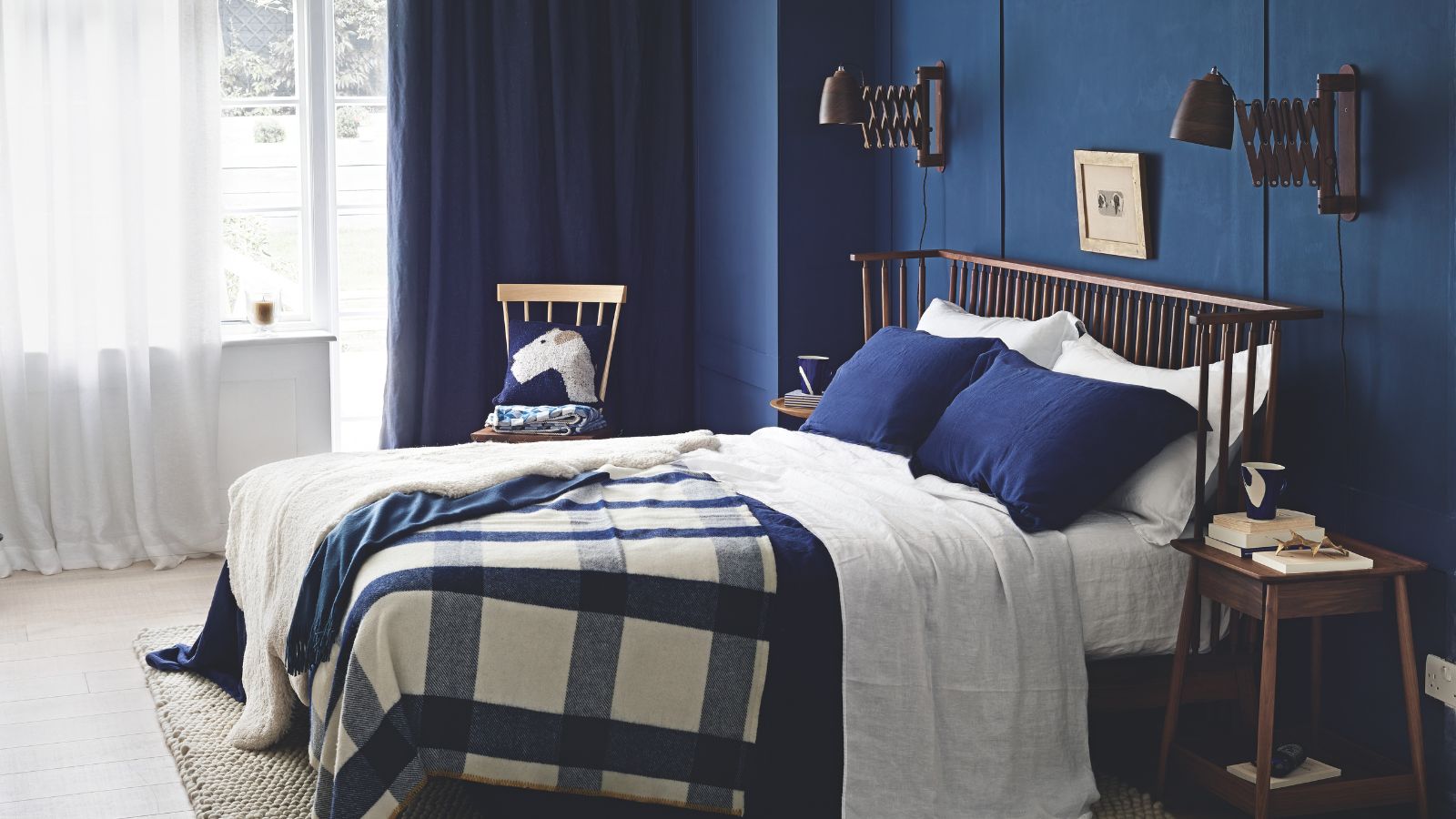 A blue bedroom with a large double bed with blue pillows and a blue and white throw, and sheer white curtains over a large window.