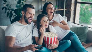 Man, woman and child watching TV together. All are smiling and eating popcorn, which the child is holding