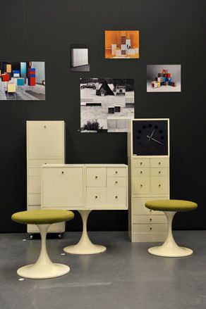 Two symmetrical shaped stools shaped like an hour glass, posiitoned infront of multiple multi-compartment white cabinets with drawers that have black handles.