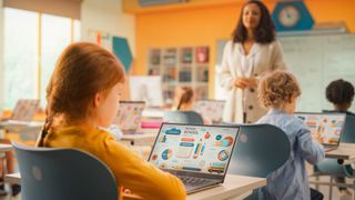 children in school on their laptops with teacher in front of class 