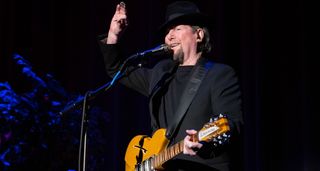 Dressed all in black, Roger McGuinn holds up his right hand and addresses the audience during a 2022 live performance. He plays a 12-string Rickenbacker.