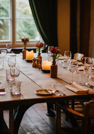 A candle-lit farmhouse wooden table is adorned with sleek glassware, ceramic flower vases, wild flowers, and silver cutlery.