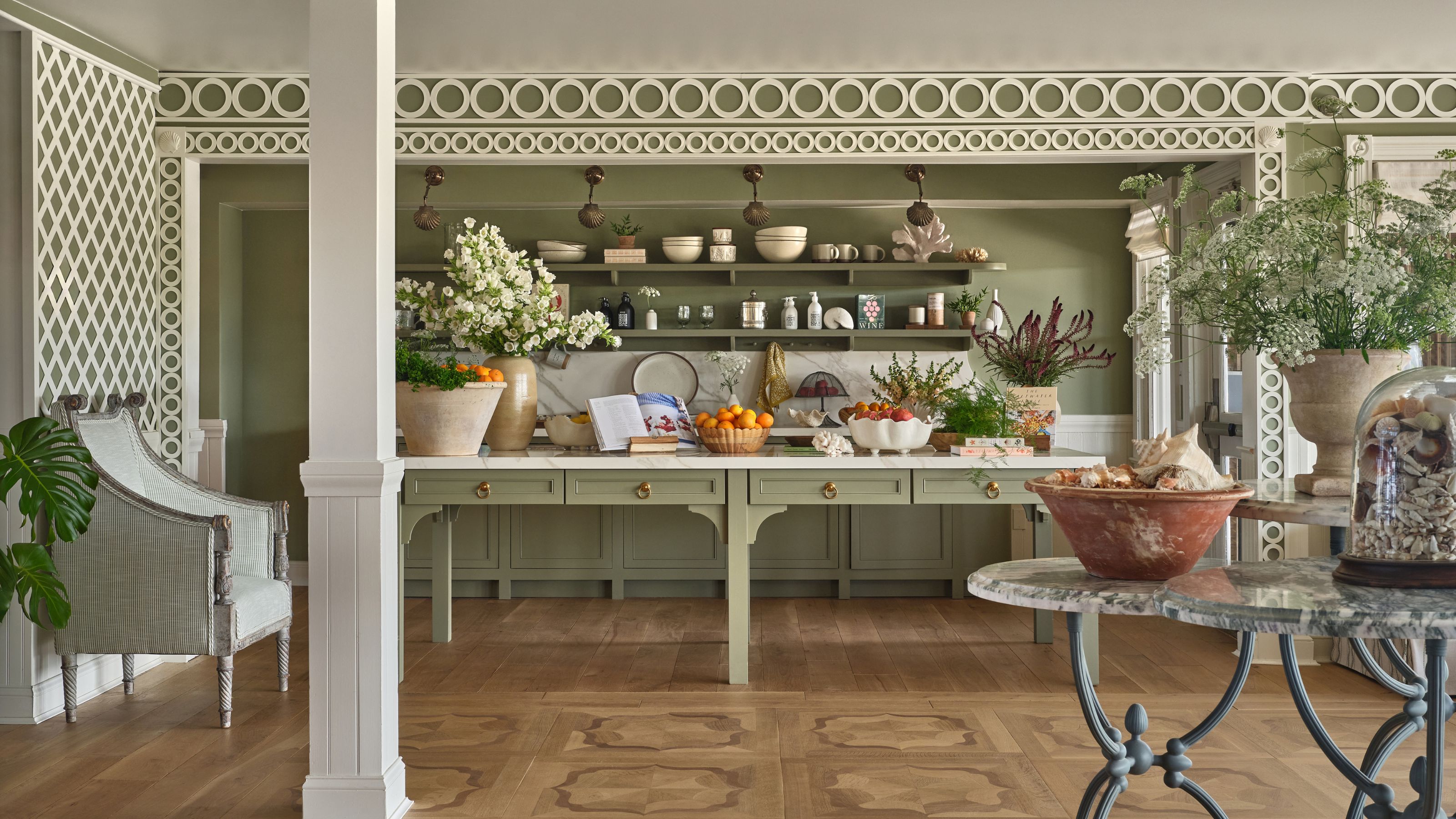 A long, sage green kitchen island topped with white marble, accented by open shelving filled with ceramics, fresh oranges, and lush white floral arrangements.